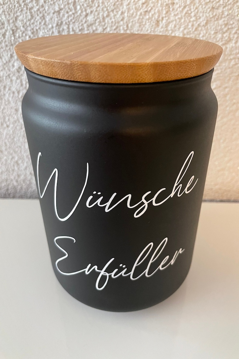 May include: Black ceramic jar with a wooden lid. The jar has white lettering that reads "W&uuml;nsche Erf&uuml;ller".