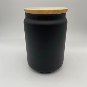 May include: A black ceramic jar with a wooden lid. The jar is cylindrical and has a matte finish.