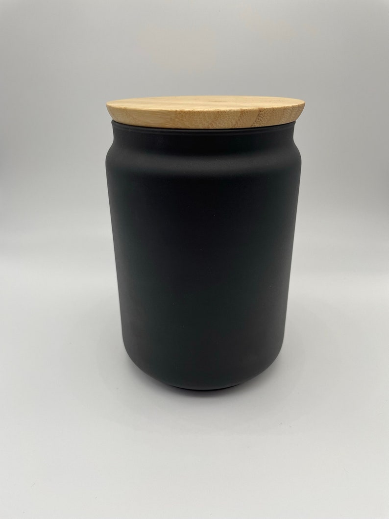 May include: A black ceramic jar with a wooden lid. The jar is cylindrical and has a matte finish.