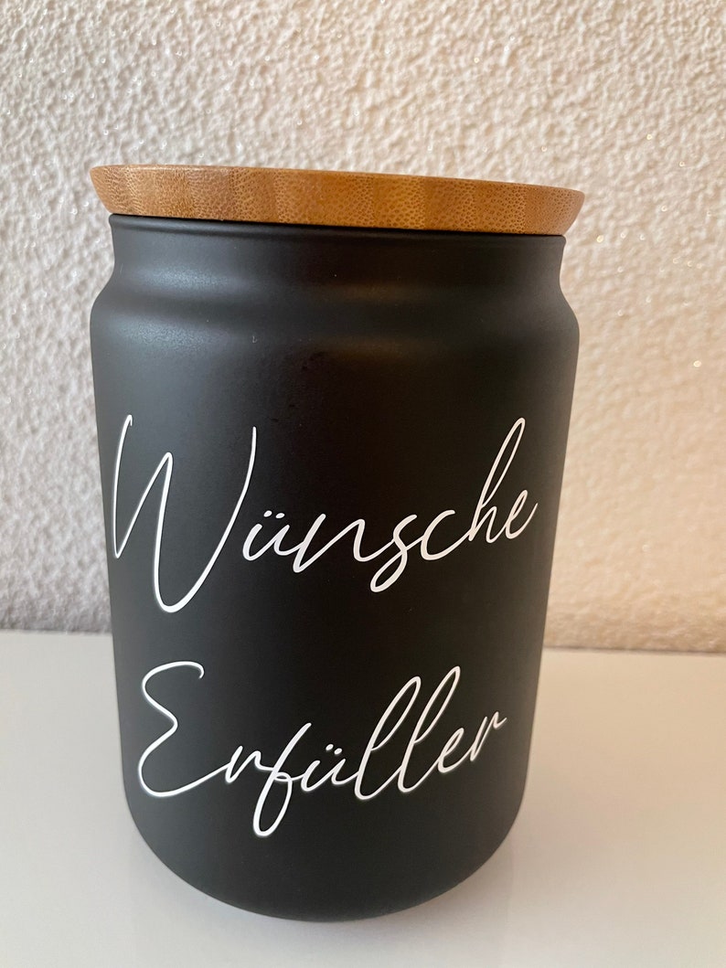 May include: Black ceramic jar with a wooden lid. The jar has white lettering that says "W&uuml;nsche Erf&uuml;ller".