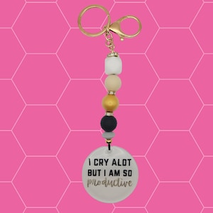 May include: A white, gold and black beaded keychain with a round white charm that says "I cry a lot but I am so productive" in black and gold glitter lettering.