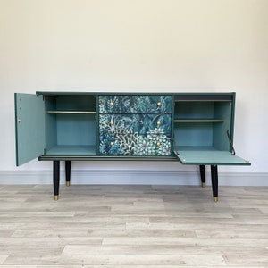 May include: A vintage-style sideboard in dark green with a botanical print on the drawers. The cabinet features two doors and a drawer, with gold-tone accents on the legs and handles. The interior is painted in a light teal colour.