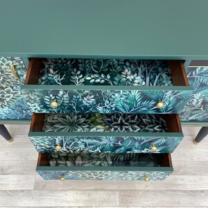 May include: A teal green dresser with three open drawers. The drawer interiors feature a lush, tropical leaf pattern in shades of green and blue. Gold-coloured knobs and black legs with gold accents complete the design.