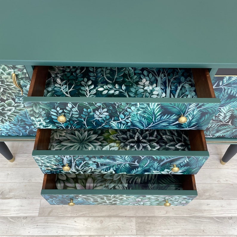 May include: A teal green dresser with three open drawers. The drawer interiors feature a lush, tropical leaf pattern in shades of green and blue. Gold-coloured knobs and black legs with gold accents complete the design.