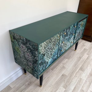 May include: A dark green sideboard with floral patterned doors and drawers. The cabinet has a solid dark green top and black legs with gold-coloured tips. The floral pattern features white, green, and blue flowers and foliage.