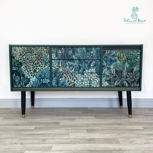 May include: A dark green sideboard with a botanical print of white and teal flowers and foliage. The cabinet has three drawers and two doors with gold hardware. The legs are black with gold tips. The text "Vibrant Wood Vintage Furniture Reimagined" is in the upper right corner.