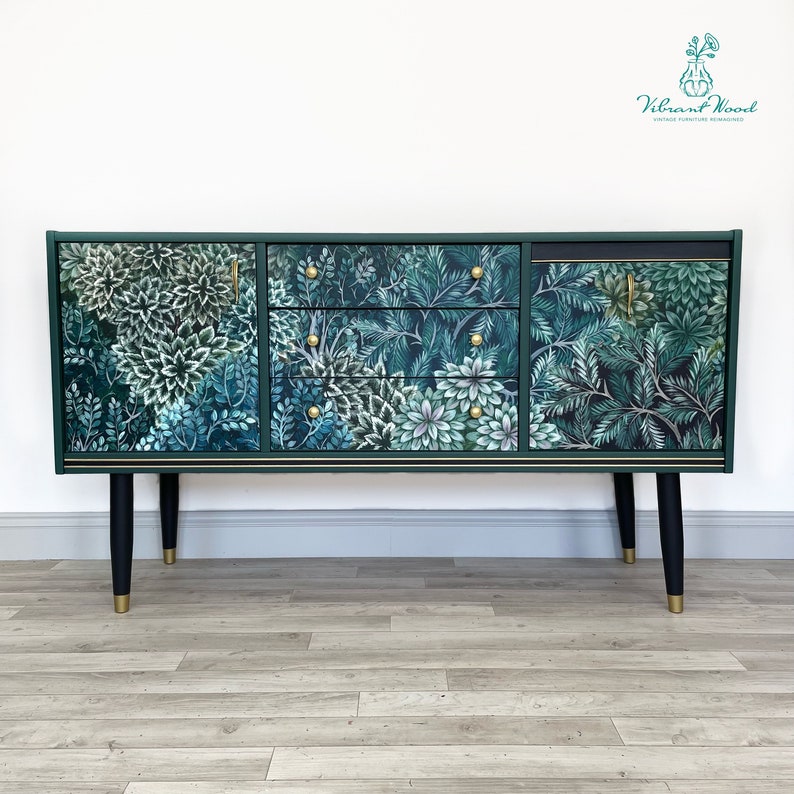 May include: A dark green sideboard with a botanical print of white and teal flowers and foliage. The cabinet has three drawers and two doors with gold hardware. The legs are black with gold tips. The text "Vibrant Wood Vintage Furniture Reimagined" is in the upper right corner.
