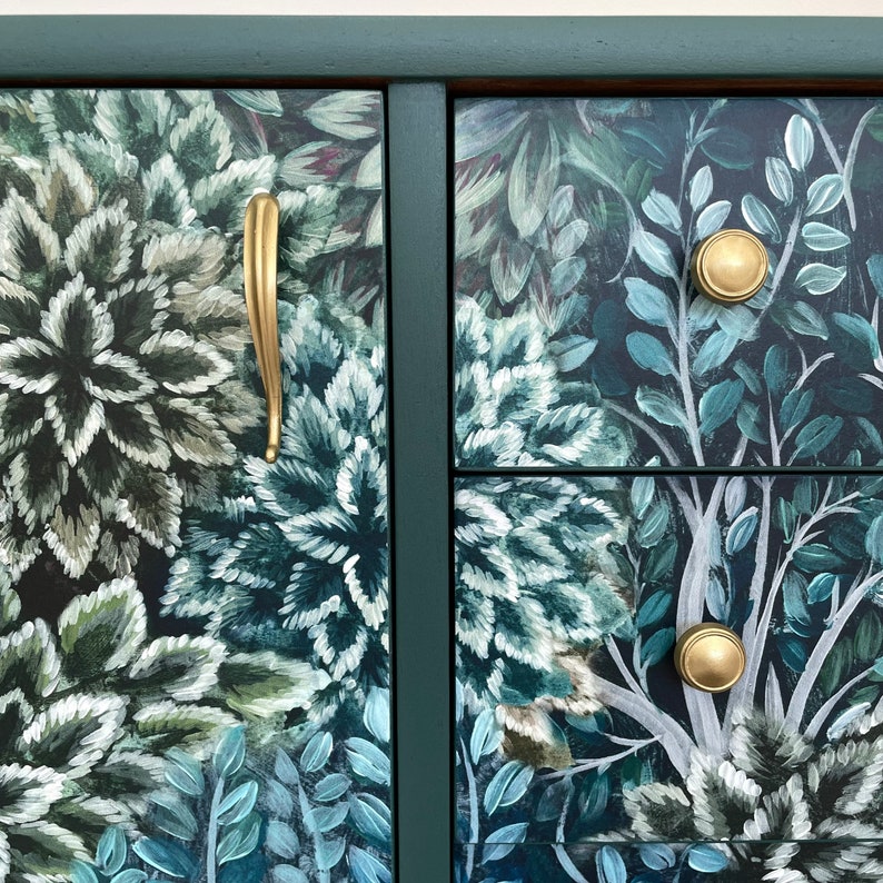 May include: A close-up of a dark green cabinet with floral artwork in shades of green, blue, and white. The cabinet has gold-coloured hardware, including a long handle and round knobs. The cabinet's frame is a darker shade of green.