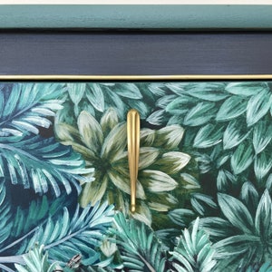 May include: Close-up of a painted cabinet drawer front with a botanical design in shades of teal, green, and white. A gold-coloured, elongated handle is centered on the drawer. The top edge is navy blue with a gold trim.
