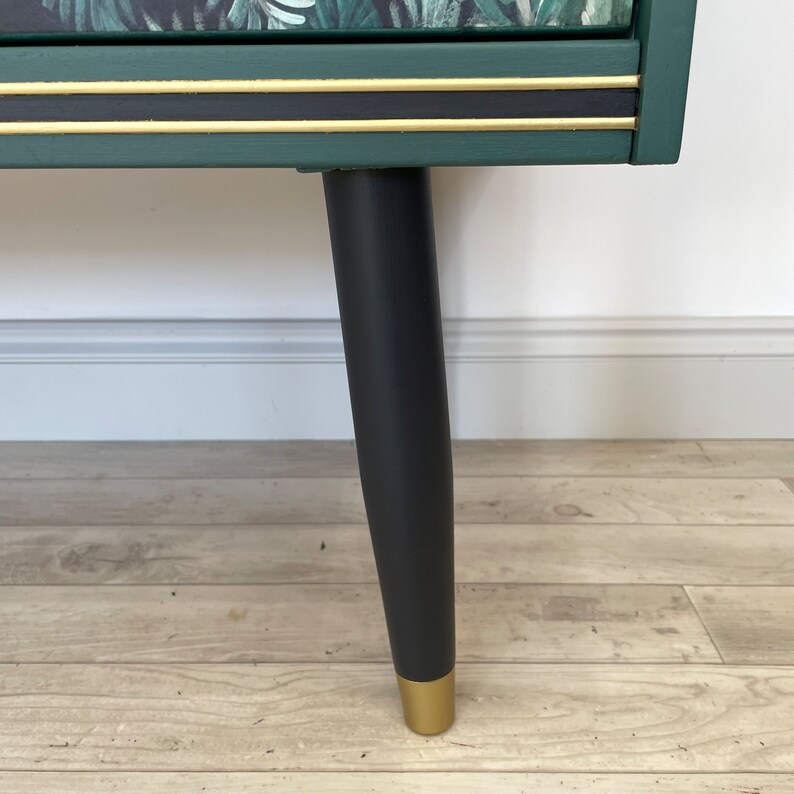 May include: A detail shot of a green cabinet with black legs and gold detailing. The cabinet has a dark green finish with gold trim and a black stripe. The legs are black with gold tips. The cabinet top has a tropical leaf design.