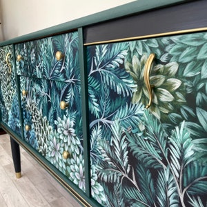 May include: A dark green sideboard with a botanical print featuring white and green foliage. The cabinet has gold hardware, including drawer pulls and handles. The legs are black with gold tips.