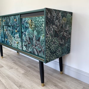 May include: A green sideboard with a botanical design in shades of green and white. It features gold handles, black legs with gold tips, and gold trim. The cabinet's surface is covered in a floral and leaf pattern, creating a natural aesthetic.