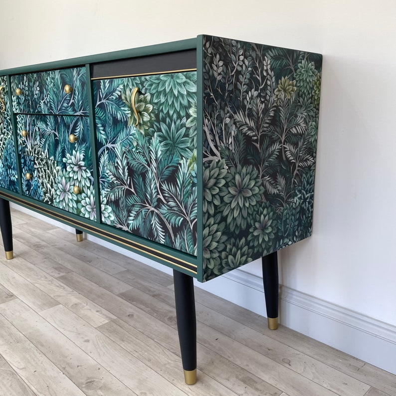 May include: A green sideboard with a botanical design in shades of green and white. It features gold handles, black legs with gold tips, and gold trim. The cabinet's surface is covered in a floral and leaf pattern, creating a natural aesthetic.