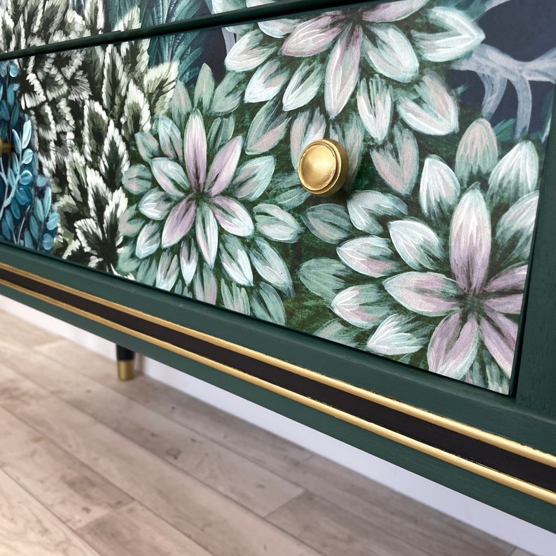 May include: A green dresser with floral drawer fronts. The drawers feature a botanical design with white, green, and lavender flowers. Gold hardware and trim accent the dark green frame. The dresser has black and gold legs.