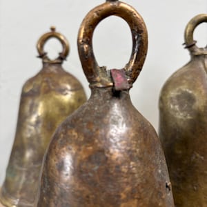 May include: Close-up of three antique bronze bells. The bells have a weathered, aged appearance with a dark brown patina. Each bell has a loop at the top for hanging. The bells are of varying sizes and show signs of wear, adding to their vintage charm.