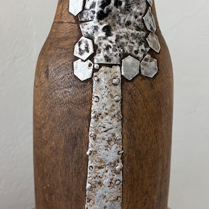 May include: A wooden vase with a dark brown finish, accented with silver metal details. The metal is applied in a vertical strip down the front and in decorative shapes around the top. The vase has a rounded shape and sits on a wooden surface.