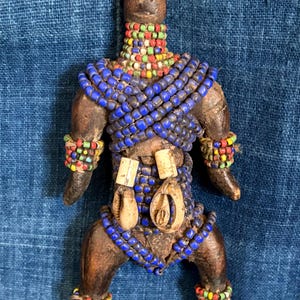 May include: A carved wooden figurine of a person, adorned with blue, red, yellow, and green beaded accents. The figure has a dark brown finish and is decorated with cowrie shells. The figure is posed on a blue fabric background.