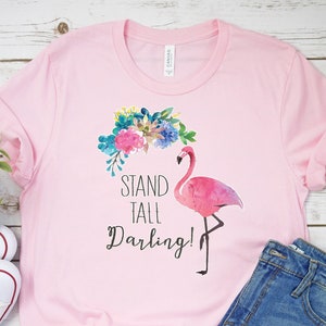 May include: A light pink t-shirt with rolled-up sleeves featuring a watercolor flamingo design and the text "Stand Tall Darling!". The flamingo is pink with a floral crown. White sneakers and denim shorts are also in the image.