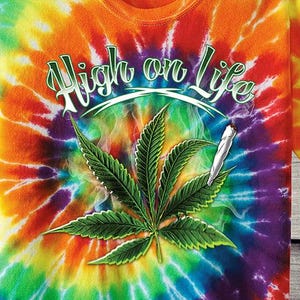 May include: A tie-dye t-shirt with a spiral rainbow pattern. The shirt features a green cannabis leaf and a lit joint, with the words "High on Life" in green script.