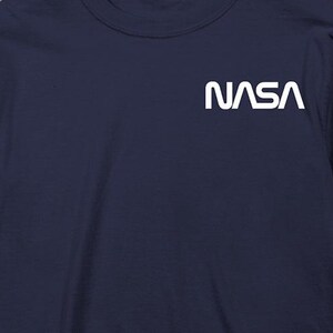 May include: Navy blue long-sleeve shirt with the white NASA logo on the upper left chest. The shirt is made of a soft material and is suitable for casual wear. The shirt is displayed on a wooden surface.