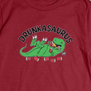 May include: A maroon t-shirt featuring a green dinosaur graphic with the text "DRUNKASAURUS". The dinosaur is lying on its back, holding a drink, with several red cups around it. The shirt is a short-sleeved crew neck.