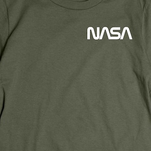 May include: Olive green t-shirt with the white NASA logo on the chest. The short-sleeved shirt is made of a soft material and is displayed on a wooden surface. A plaid shirt and a pair of jeans are partially visible.