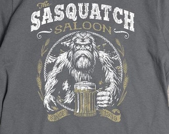 Puede incluir: Una camiseta gris oscuro con un gráfico blanco de un sasquatch sosteniendo una jarra de cerveza. El gráfico incluye el texto "The Sasquatch Saloon" y "Est. 1978".