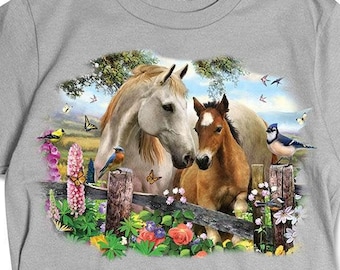 Può includere: T-shirt grigio chiaro con una grafica dettagliata di due cavalli, una giumenta bianca e un puledro marrone, in piedi dietro una staccionata di legno. La scena comprende fiori colorati, farfalle e uccelli, sullo sfondo di alberi e cielo blu.