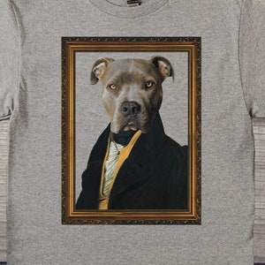 May include: A gray t-shirt featuring a framed portrait of a dog dressed in formal attire. The dog has a gray coat and is wearing a black suit jacket with a yellow vest and a black bow tie. The portrait is in a gold frame.