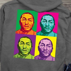 May include: Charcoal gray hoodie with a front pocket and drawstring hood. The front features a small white graphic of three faces with the text "THE THREE STOOGES". The back displays a pop art style image of a face in four different color blocks.