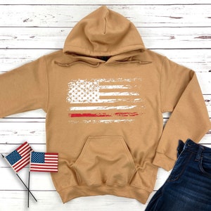 May include: A tan hoodie with a distressed white American flag and a red stripe across the middle. The hoodie has a kangaroo pocket and a drawstring hood.