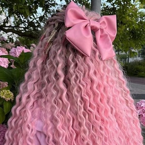 May include: Long, wavy, pastel pink hair extensions featuring a large, matching pink satin hair bow attached at the crown. The hair has a crimped, textured appearance with some braided sections integrated throughout the length of the hair.
