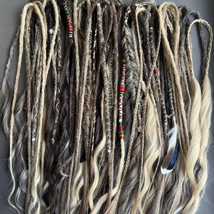 May include: A collection of synthetic hair extensions featuring a mix of blonde, brown, and grey tones. The set includes various textures such as dreadlocks, braids, and loose wavy strands, accented with decorative beads, thread wraps, and a single feather.