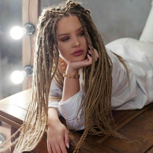 Op de afbeelding: Lange, lichtbruine synthetische dreadlocks in een natuurlijke, getextureerde stijl. De haarextensies hebben een meerkleurig uiterlijk met variërende diktes in elke streng, wat zorgt voor een volumineus en gelaagd effect rondom het gezicht.