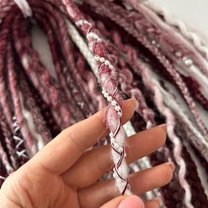 May include: Close-up of burgundy and white dreadlocks. One dreadlock is decorated with white beads and burgundy and white thread. The dreadlocks are tied together with a white band.