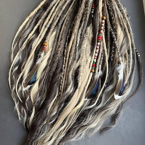 May include: A set of synthetic hair extensions featuring a blend of dark brown, light brown, and blonde strands. The collection includes various textures, including braided sections, wrapped cords, wooden beads, and three feathers with blue tips.