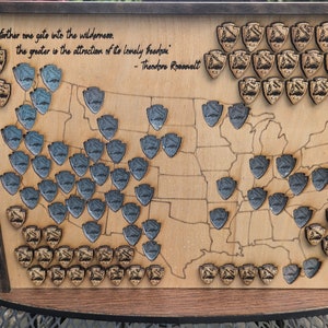 May include: Wooden map of the United States with engraved state outlines. The map features numerous shield-shaped pins in gold and blue, marking locations. A quote by Theodore Roosevelt is inscribed at the top.