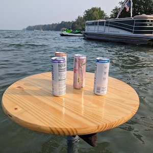 May include: Four cans of seltzer on a round, wooden table floating in water. The cans are silver, pink, and white with colorful labels. A pontoon boat and jet ski are in the background. The table is supported by a metal pole.