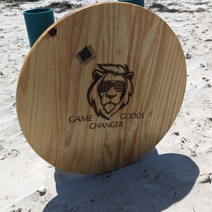 May include: A round wooden table with two teal blue cup holders. The table has a lion with American flag sunglasses and the text "GAME CHANGER GOODS" burned into the wood.