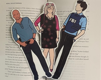 May include: Three illustrated bookmarks featuring characters from a book. The bookmarks are printed on white cardstock and feature a woman with short blonde hair wearing a pink and black dress, a man with dark hair wearing a blue shirt and black pants, and a man with dark hair wearing a blue shirt and black pants with an FBI badge.