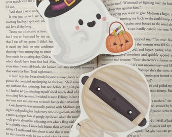 Puede incluir: Tres recortes de papel con temática de Halloween. Uno es un fantasma con un sombrero de bruja y una cesta de calabaza. Otro es una momia. El tercero es un fantasma con una cesta de calabaza. Todos están en una página de libro.
