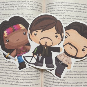 May include: Three illustrated bookmarks featuring characters from the TV show The Walking Dead. The bookmarks are in a chibi style, with large heads and small bodies. The characters are Daryl Dixon, Michonne, and Rick Grimes. Daryl is holding a crossbow, Michonne is holding a katana, and Rick is holding a pistol. The bookmarks are printed on cardstock and have a matte finish.