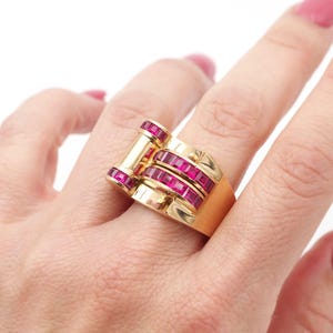 Peut inclure: Une bague en or avec un design rectangulaire, ornée de plusieurs rangées de pierres précieuses rouges taillées en carré. La bague est posée sur un fond blanc, mettant en valeur ses détails complexes et sa couleur vibrante. La bague est sur un doigt.