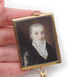 Peut inclure: Un portrait miniature d'un homme dans un cadre doré. Le portrait représente un homme à la peau pâle, aux yeux bleus et aux cheveux blancs, portant un manteau noir et une chemise blanche. Un petit cadenas doré est suspendu au bas du cadre.