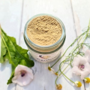 May include: A glass jar filled with a light brown powder labelled "New Moon Digestive Herbal Tea Blend". The jar is sitting on a white wooden surface with green and white flowers.