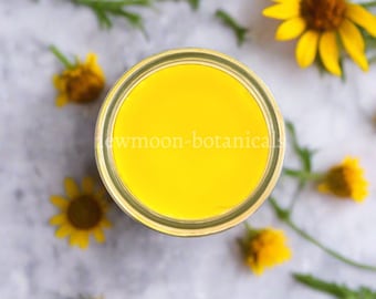 Könnte beinhalten: Draufsicht auf ein Glas mit leuchtend gelber Flüssigkeit, möglicherweise ein Kosmetikprodukt, mit dem Text "newmoon-botanicals" auf dem Glas. Das Glas ist von gelben Blumen und grünem Laub auf einer hellgrauen Oberfläche umgeben.