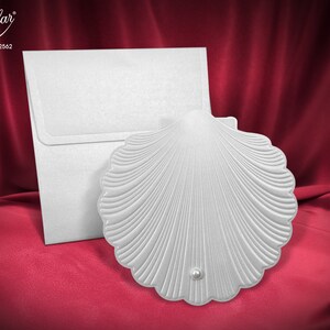 Puede incluir: Tarjeta de invitación blanca en forma de concha de vieira con un acento de perla. La tarjeta está sobre un fondo rojo y tiene el texto "Popular Collection - 2562" impreso en ella.