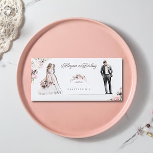Può includere: Un biglietto d'invito a nozze con i nomi "Aleyna ve Berkay" e la data "26.07.24" stampati. Il biglietto presenta illustrazioni di una sposa e uno sposo con accenti floreali. Il biglietto è su un piatto rosa.