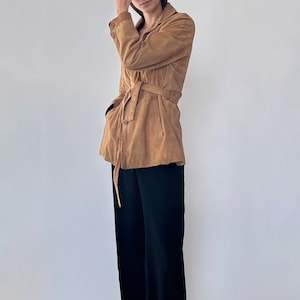 May include: A brown suede jacket with a belt and long sleeves. The jacket is worn over black pants and white sneakers.