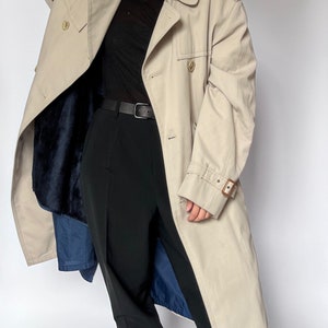 May include: A beige trench coat with a brown leather belt and a blue lining. The coat has a double-breasted closure and a collar.